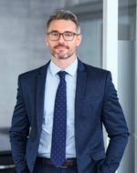Portrait of handsome mid adult business man standing in modern office. Successful mature entrepreneur in formal clothing looking at camera with satisfaction. Confident man in suit with eyeglasses and beard standing with hands in pocket and looking at camera in office.